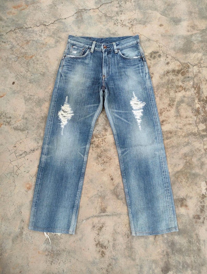 May include: Light blue denim jeans with distressed details, including ripped sections on the front of the legs. The jeans have a classic five-pocket design and a button and zipper closure. The denim has a faded wash.