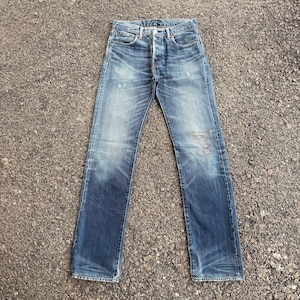 Puede incluir: Vaqueros de mezclilla azul descolorido con bragueta de botones. Los vaqueros muestran signos de desgaste, con parches más claros y decoloración en toda la tela. Los vaqueros están colocados planos sobre una superficie texturizada.