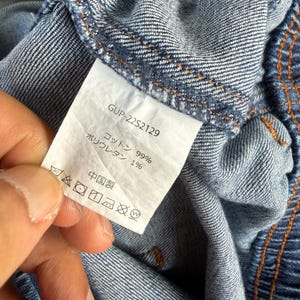 May include: Close-up of blue denim fabric with a white tag. The tag has the text "GUP-22S2129", and Japanese text indicating the fabric composition: 99% cotton and 1% polyurethane. The tag also includes care symbols.