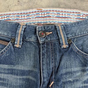 May include: Close-up of blue denim jeans with a decorative patterned waistband in shades of blue, red, and white. The jeans feature a button closure, belt loops, and detailed stitching. The pockets have brown leather accents.
