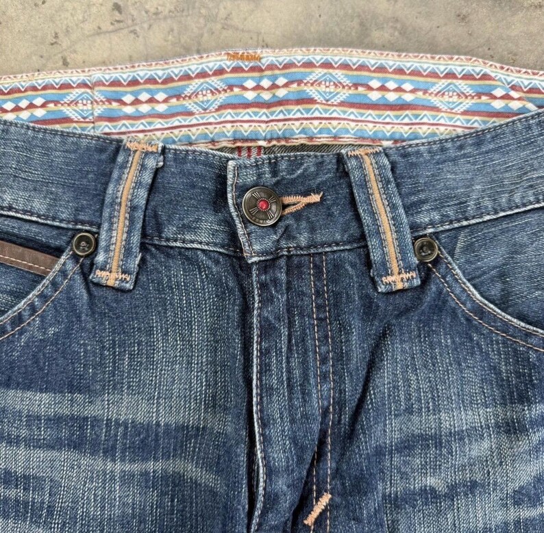 May include: Close-up of blue denim jeans with a decorative patterned waistband in shades of blue, red, and white. The jeans feature a button closure, belt loops, and detailed stitching. The pockets have brown leather accents.