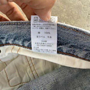 Pu&ograve; includere: Primo piano dell'interno di un capo in denim, che mostra un'etichetta bianca con testo giapponese e istruzioni per la cura. L'etichetta indica 100% cotone e un'etichetta in pelle. Il tessuto denim &egrave; un classico lavaggio blu.