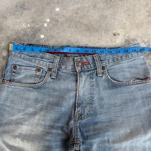 May include: Light blue denim jeans with a red accent on the waistband and pockets. A blue measuring tape is placed across the waist, showing a measurement of approximately 17 inches.