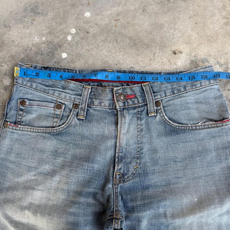 May include: Light blue denim jeans with a red accent on the waistband and pockets. A blue measuring tape is placed across the waist, showing a measurement of approximately 17 inches.