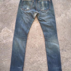 May include: Faded blue denim jeans with a worn appearance. The jeans have a classic five-pocket design, a leather patch, and visible wear and tear on the back pockets and knees. The jeans are displayed on a neutral surface.