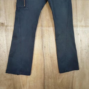Pu&ograve; includere: Pantaloni blu navy scuro con tasca cargo e dettaglio zip. I pantaloni hanno un design a gamba dritta e sono realizzati in un tessuto resistente. I pantaloni sono mostrati su uno sfondo di legno marrone chiaro.