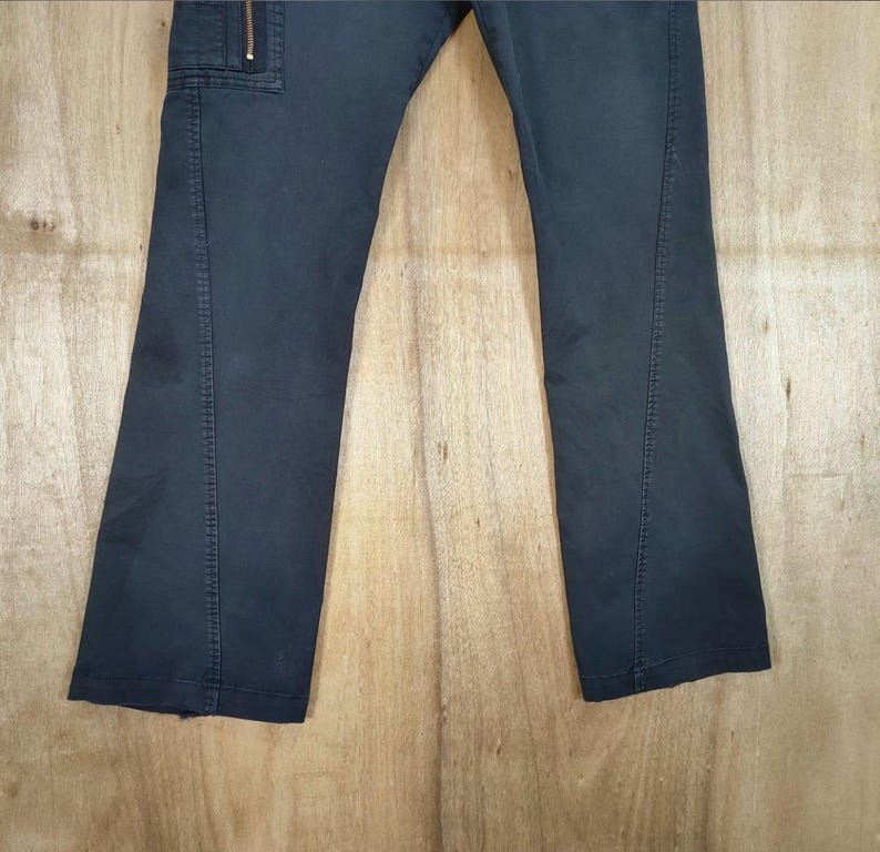 Pu&ograve; includere: Pantaloni blu navy scuro con tasca cargo e dettaglio zip. I pantaloni hanno un design a gamba dritta e sono realizzati in un tessuto resistente. I pantaloni sono mostrati su uno sfondo di legno marrone chiaro.