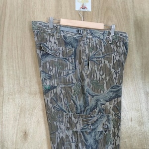 May include: Camouflage cargo trousers with multiple pockets and button fastenings. The trousers feature a tree bark pattern in shades of brown, green, and grey. The trousers are hanging on a wooden hanger.