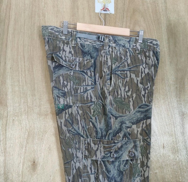 May include: Camouflage cargo trousers with multiple pockets and button fastenings. The trousers feature a tree bark pattern in shades of brown, green, and grey. The trousers are hanging on a wooden hanger.