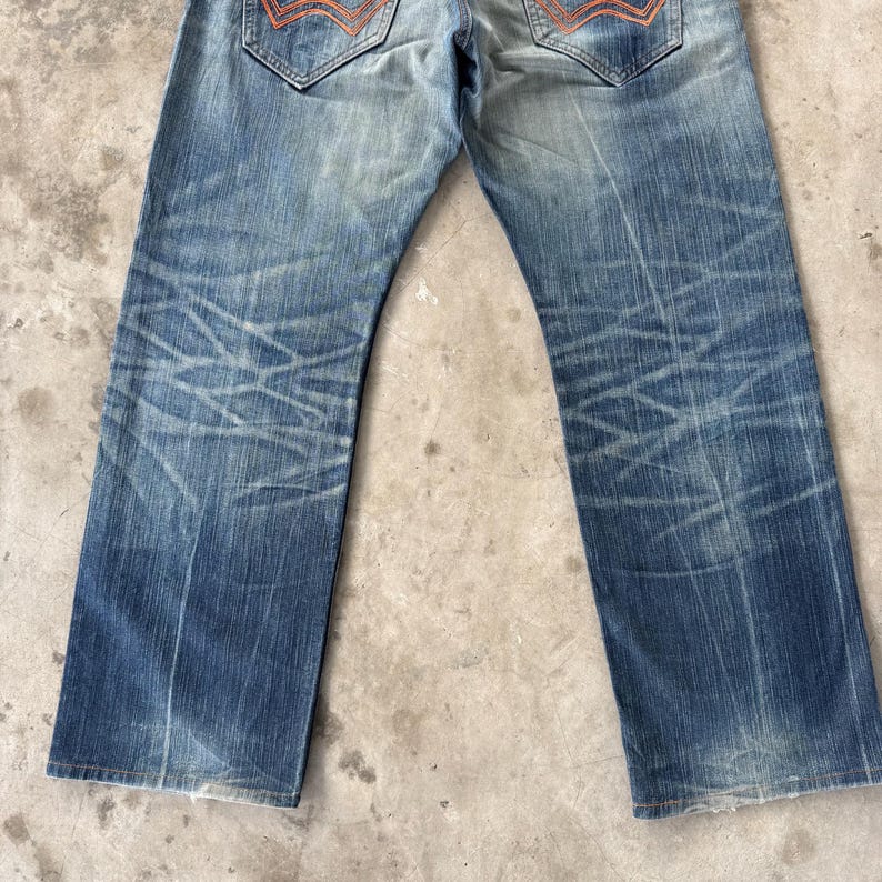 Pu&ograve; includere: Jeans in denim blu sbiadito con dettagli distressed. I jeans presentano un classico design a cinque tasche, con cuciture arancioni sulle tasche posteriori. Il denim ha un aspetto usurato con aree sbiadite e sottili baffature.