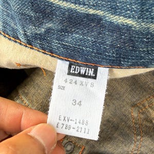 Pu&ograve; includere: Primo piano di un'etichetta di abbigliamento in denim con il marchio "EDWIN" stampato in bianco. L'etichetta indica la taglia "34" e include codici prodotto. Il tessuto denim &egrave; blu scuro con cuciture visibili.
