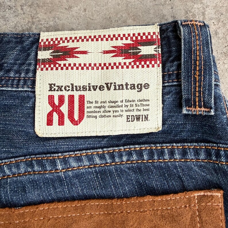 Op de afbeelding: Close-up van een blauwe spijkerbroek met een bruine leren patch. De jeans heeft een rechthoekig label met de tekst "Exclusive Vintage" en "XU". Het label bevat ook de tekst "EDWIN".