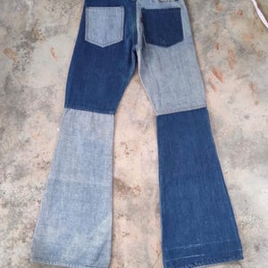 May include: Two-tone denim jeans with a patchwork design. The jeans feature a light blue and dark blue color scheme, with a flared leg. The back pockets are visible, and the jeans are laid flat on a concrete surface.