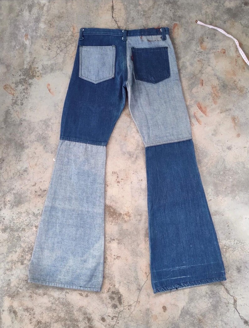 May include: Two-tone denim jeans with a patchwork design. The jeans feature a light blue and dark blue color scheme, with a flared leg. The back pockets are visible, and the jeans are laid flat on a concrete surface.