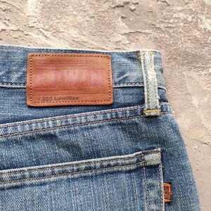 May include: Close-up of blue denim jeans with a brown leather patch. The patch features the brand name "Edwin" in a faded pink color, with "505 Lowrise" printed below. An orange "Edwin" tag is visible on the back pocket.