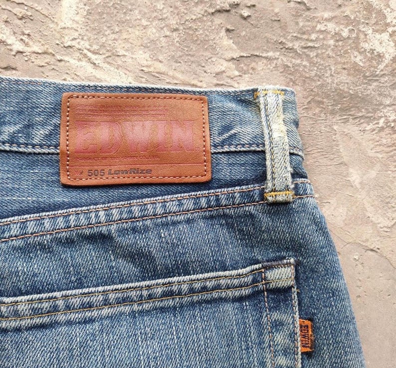 May include: Close-up of blue denim jeans with a brown leather patch. The patch features the brand name "Edwin" in a faded pink color, with "505 Lowrise" printed below. An orange "Edwin" tag is visible on the back pocket.