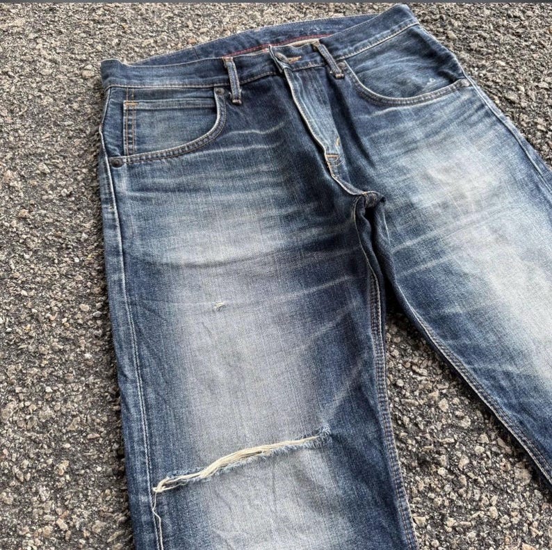 Pu&ograve; includere: Primo piano di jeans blu con un aspetto usurato e vissuto. I jeans hanno un lavaggio sbiadito, cuciture visibili e uno strappo sul ginocchio. Il tessuto mostra segni di usura, conferendo un aspetto vintage.
