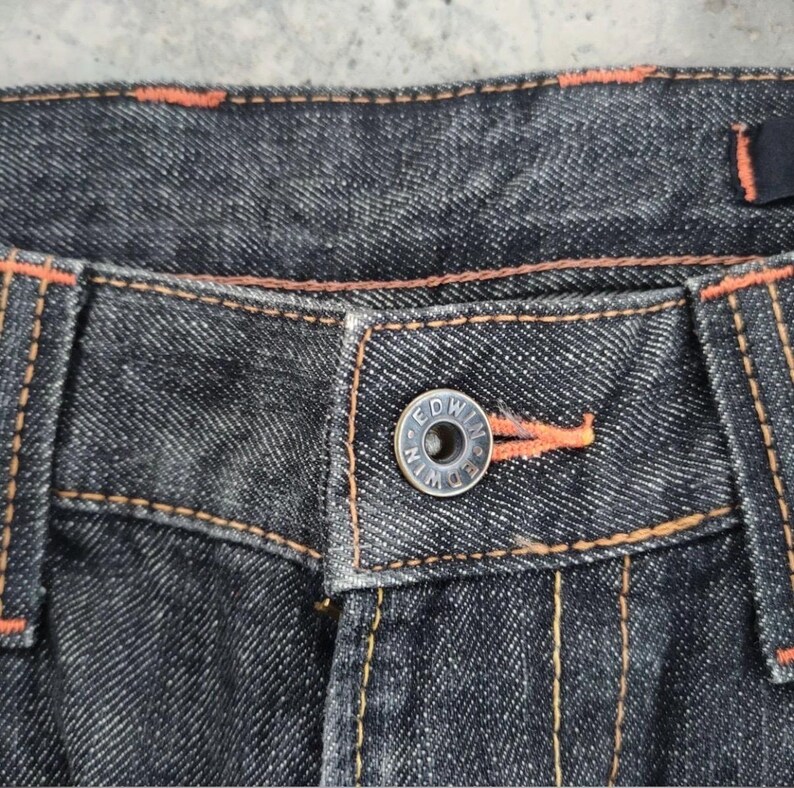 Pu&ograve; includere: Primo piano di jeans di denim scuro con cuciture arancioni. I jeans presentano un bottone argentato con il marchio "EDWIN" e dettagli di filo arancione sulla patta. Il tessuto ha un aspetto strutturato.