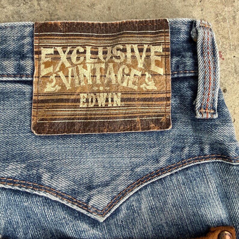May include: Close-up of blue denim jeans with a brown leather patch. The patch features the text "EXCLUSIVE VINTAGE" and "EDWIN" in a vintage style. The jeans have a classic five-pocket design with a stitched back pocket.