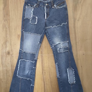 Puede incluir: Pantalones vaqueros azules con diseño de parches, bordes deshilachados y costuras expuestas. Los pantalones tienen cintura de tiro medio, cierre de botón y cremallera, y pernera acampanada. El tejido denim muestra diversas tonalidades de azul.