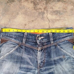 以下が含まれることがあります： ウエストに黄色のメジャーを当てた、青いデニムジーンズのクローズアップ。ジーンズには茶色の革のアクセントとボタン留めが付いています。ウエストは約46cmです。