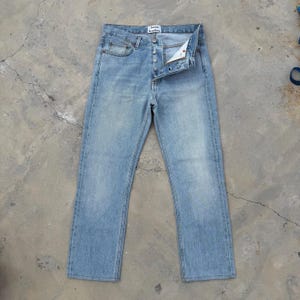 May include: Light blue denim jeans with a button fly and five-pocket design. The jeans have a faded wash and a straight leg fit. The brand name "Acne Studios" is visible on the back waistband.