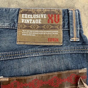 May include: Close-up of blue denim jeans with a brown leather patch. The patch features the text "EXCLUSIVE VINTAGE" and "EDWIN". The jeans have a classic five-pocket design and a button and zipper closure.