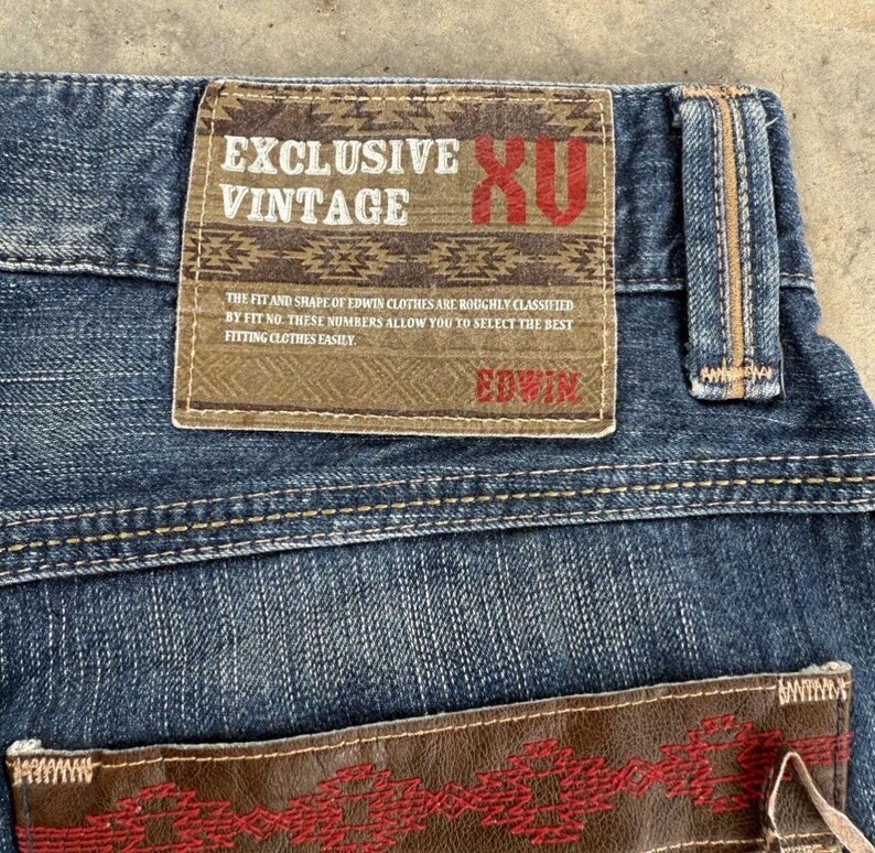 May include: Close-up of blue denim jeans with a brown leather patch. The patch features the text "EXCLUSIVE VINTAGE" and "EDWIN". The jeans have a classic five-pocket design and a button and zipper closure.