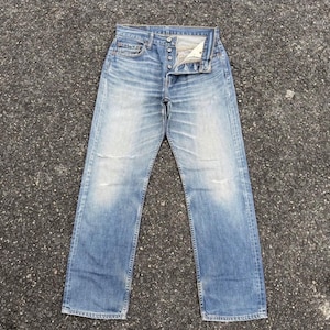 Könnte beinhalten: Verblasste blaue Jeans mit Knopfleiste. Die Jeans haben ein klassisches Fünf-Taschen-Design und weisen Gebrauchsspuren auf, einschließlich abgenutzter Bereiche an den Knien. Die Jeans liegen flach auf einer strukturierten Oberfläche.