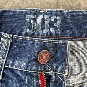 May include: Close-up of a pair of blue denim jeans. The jeans feature the number "503" printed on the waistband. A copper-colored button is visible, along with a red zipper.