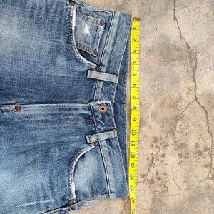 May include: Close-up of blue denim jeans with a distressed pocket and a yellow measuring tape. The jeans have a button closure and belt loops. The measuring tape shows a measurement of approximately 16 inches.