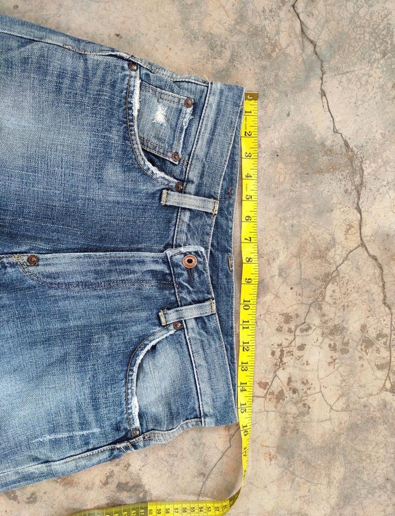 May include: Close-up of blue denim jeans with a distressed pocket and a yellow measuring tape. The jeans have a button closure and belt loops. The measuring tape shows a measurement of approximately 16 inches.