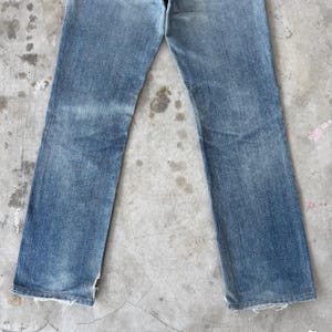 May include: Light blue denim jeans with a straight leg cut. The jeans show signs of wear with faded areas and frayed edges at the bottom of the legs. The jeans are laid flat on a concrete surface.