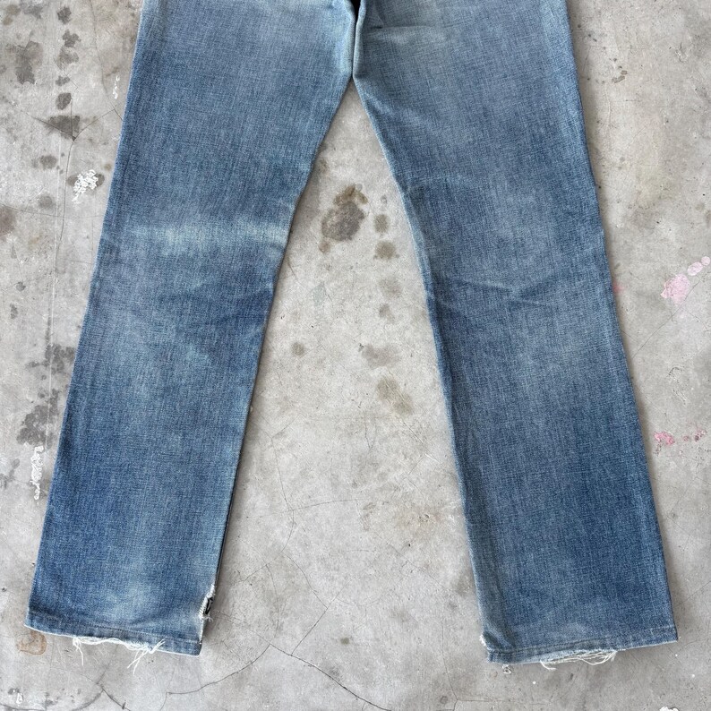 May include: Light blue denim jeans with a straight leg cut. The jeans show signs of wear with faded areas and frayed edges at the bottom of the legs. The jeans are laid flat on a concrete surface.