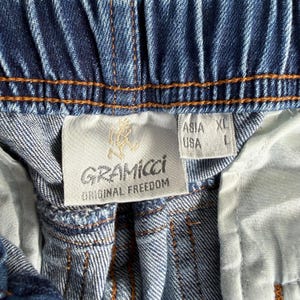 May include: Close-up of blue denim shorts with an elastic waistband and orange stitching. A white tag displays the brand name "Gramicci" and "Original Freedom" with a logo. The tag also indicates size XL for Asia and L for the USA.