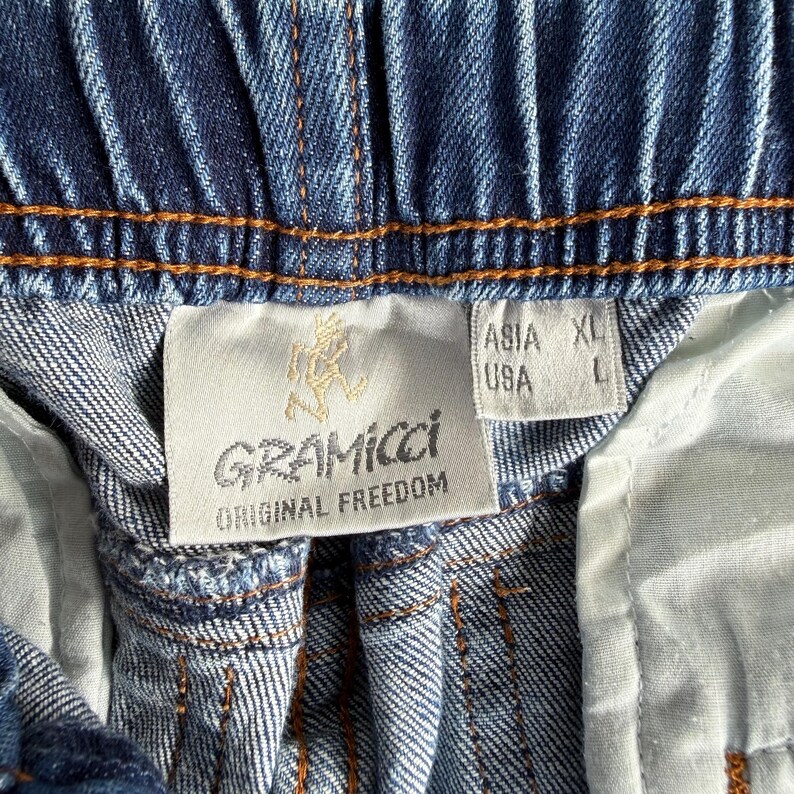May include: Close-up of blue denim shorts with an elastic waistband and orange stitching. A white tag displays the brand name "Gramicci" and "Original Freedom" with a logo. The tag also indicates size XL for Asia and L for the USA.