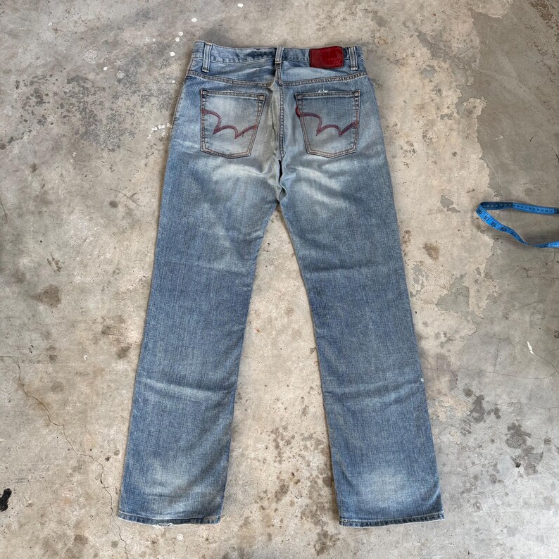 May include: Light blue denim jeans with a faded wash. The back pockets feature a unique red stitching design. A red leather patch is visible at the waistband. The jeans are laid flat on a concrete surface.