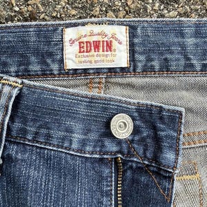 May include: Close-up of blue denim jeans with a button and zipper. A rectangular label reads "Genuine Quality Jeans EDWIN Exclusive design for lasting good look."