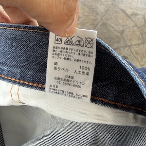 May include: A close-up of a tag attached to denim fabric. The tag has Japanese text and care symbols, including a 40-degree wash symbol. The tag also indicates the material composition: 100% cotton and artificial leather.