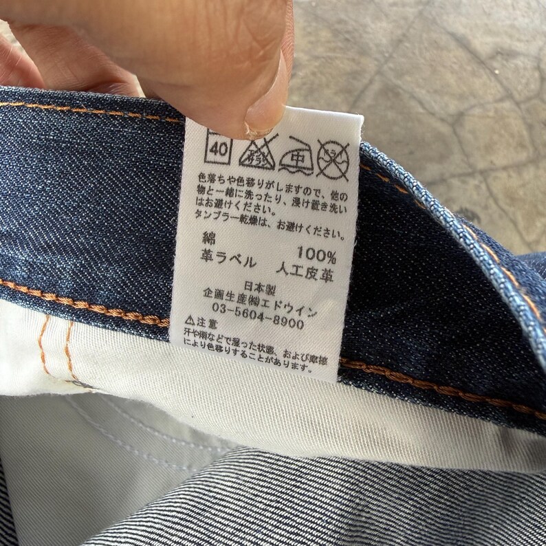 May include: A close-up of a tag attached to denim fabric. The tag has Japanese text and care symbols, including a 40-degree wash symbol. The tag also indicates the material composition: 100% cotton and artificial leather.