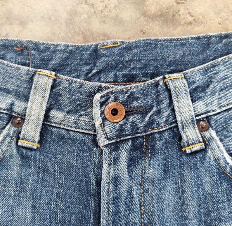 May include: Close-up of blue denim jeans. The jeans feature a copper-colored button and rivets, with yellow stitching details. The fabric shows signs of wear, with a faded appearance.
