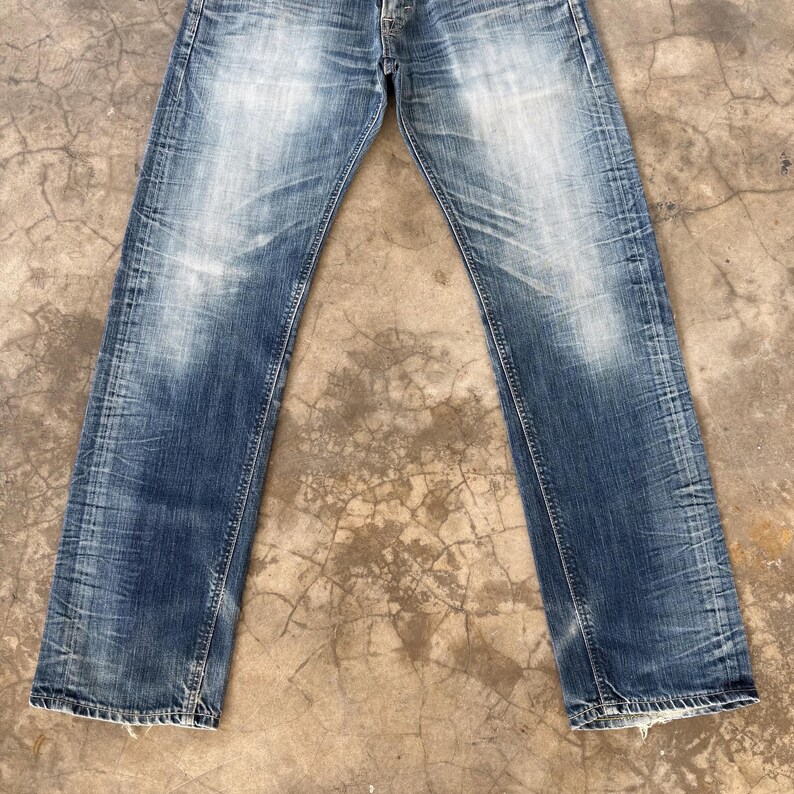 May include: Faded blue denim jeans with a straight leg cut. The jeans show signs of wear with lighter patches and creases throughout the fabric. The jeans are button-fly and have classic five-pocket styling.