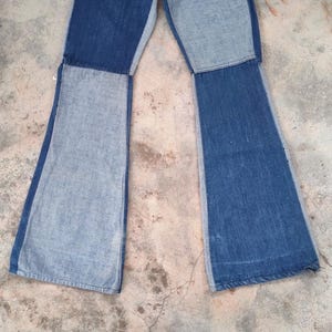 May include: A pair of blue denim bell-bottom jeans with a patchwork design. The jeans feature a light blue denim panel on the sides and a darker blue denim panel in the center of each leg. The jeans are laid flat.