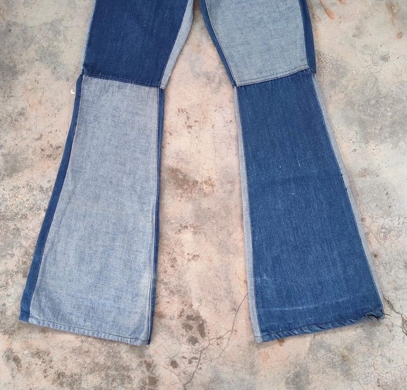 May include: A pair of blue denim bell-bottom jeans with a patchwork design. The jeans feature a light blue denim panel on the sides and a darker blue denim panel in the center of each leg. The jeans are laid flat.
