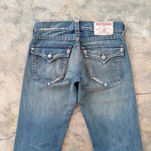 May include: Faded blue denim jeans with decorative stitching on the back pockets and a True Religion label. The jeans have a classic five-pocket design and a button and zip closure. The jeans are a classic style.