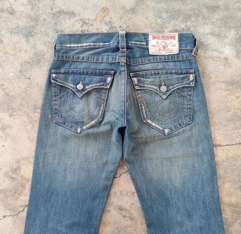 May include: Faded blue denim jeans with decorative stitching on the back pockets and a True Religion label. The jeans have a classic five-pocket design and a button and zip closure. The jeans are a classic style.