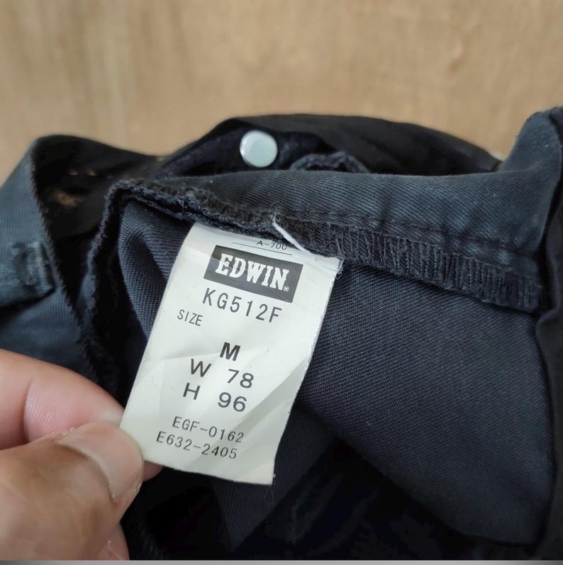 Pu&ograve; includere: Primo piano di jeans neri con un'etichetta bianca. L'etichetta recita "EDWIN" e include la taglia "M", la misura della vita "W 78 cm" e la misura dell'altezza "H 96 cm". I jeans sono in tessuto scuro.