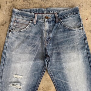 May include: Light blue denim jeans with a faded wash and distressed details. The jeans feature a classic five-pocket design, a button and zipper closure, and visible wear and tear on the front.