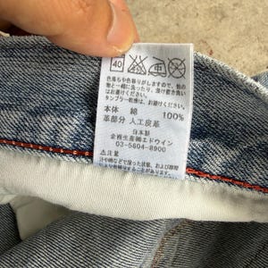 May include: Close-up of a denim garment label with Japanese text. The label includes washing instructions, material composition (100% cotton), and the manufacturer's information. The label is attached to a blue denim fabric.