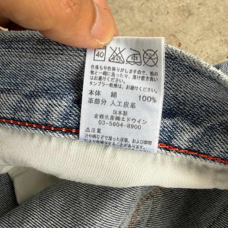 May include: Close-up of a denim garment label with Japanese text. The label includes washing instructions, material composition (100% cotton), and the manufacturer's information. The label is attached to a blue denim fabric.